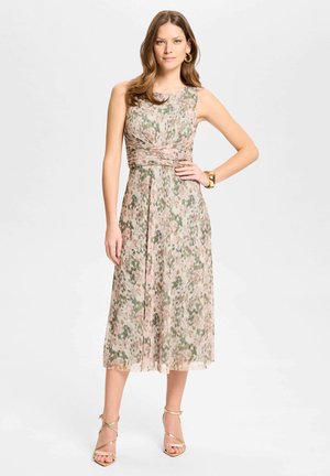 Frau trägt ein ärmelloses, knielanges Blumen-Kleid in Grün- und Pinktönen, kombiniert mit goldenen Sandalen, Creolen und einem goldenen Armband.