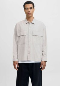 Camicia grigia chiara con motivo a griglia, due tasche sul petto e una vestibilità rilassata. Indossata sopra una camicia bianca e pantaloni neri.
