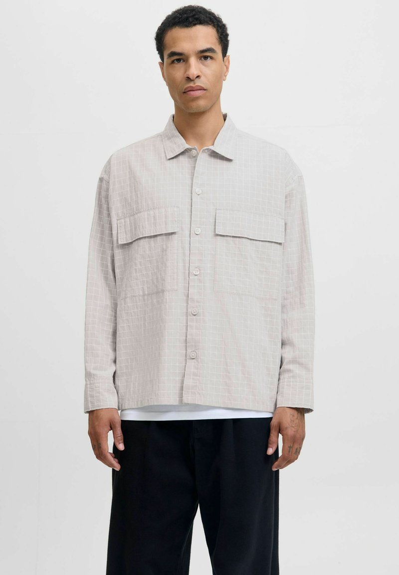 Camicia grigia chiara con motivo a griglia, due tasche sul petto e una vestibilità rilassata. Indossata sopra una camicia bianca e pantaloni neri.