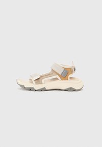 Beige outdoor sandal with adjustable straps, textured sole, and cushioned footbed. Features gray accents and a rugged design for grip.