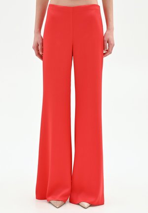 Femme portant un pantalon taille haute, large et rouge vif, associé à des talons pointus beige sur fond blanc.