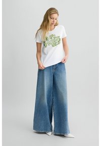 T-shirt en coton blanc avec un motif floral vert, associé à un jean en denim bleu à jambes larges. Le tissu semble doux avec une coupe décontractée et droite.