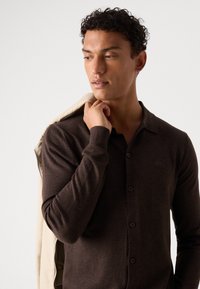Bruine gebreide polo shirt met een knoopsluiting aan de voorkant en lange mouwen, met een zachte textuur; een lichte jas over de schouder gedrapeerd.