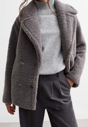 Kvinde iført en grå, blød dobbeltknappet frakke, lys grå ribstrikket rullekravesweater og mørkegrå plisserede bukser med hånden i lommen.