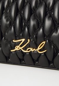 Svart quilted skinnveske med en gullfarget metal "Karl"-logotype. Overflaten har en teksturert finish med fremtredende polstrede seksjoner.