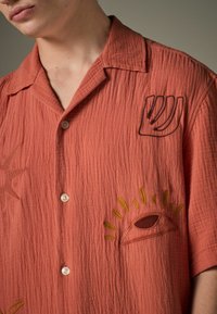Rustrød skjorte med struktureret stof, prydet med broderede sol- og øje-designs samt en button-down krave. Hvide knapper.