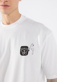 White cotton t-shirt featuring a black graphic design of a butterfly and wireless earbuds on the left chest area.