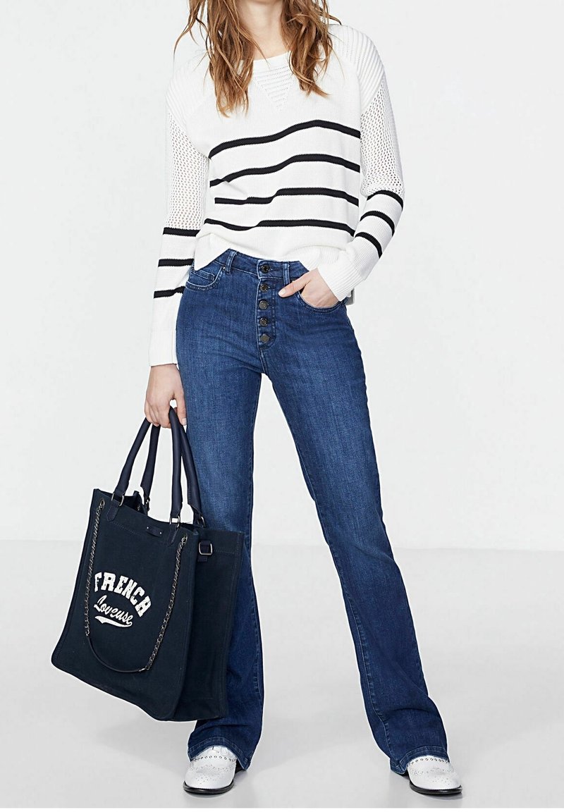 Pull en maille blanche avec des rayures noires, jeans évasés bleus, et un sac fourre-tout bleu marine avec des lettres blanches. Chaussures blanches avec un détail accentué au niveau de l’orteil.