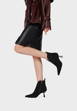 Gambe con stivaletti neri a punta con tacco basso, gonna nera in pelle e giacca marrone scuro con cintura su sfondo chiaro.