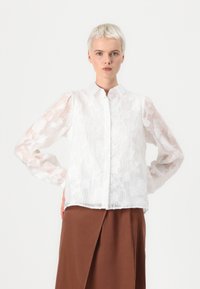 Hvit skjorte med blomsterbroderi, knappelukking foran og gjennomsiktige lange ermer. Strukturert stoff med en litt løs passform.