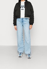 Cazadora bomber negra cortada, camiseta gráfica gris con "NYC", pantalones vaqueros de pierna ancha azul claro con slit en los laterales, y zapatillas blancas con detalles azules.