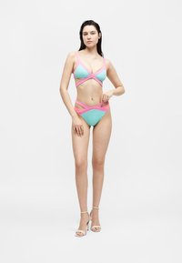 Γυναίκα που στέκεται φορώντας μπλε και ροζ μαγιό με κοψίματα και λευκά σανδάλια με ψηλό τακούνι σε απλό λευκό φόντο.