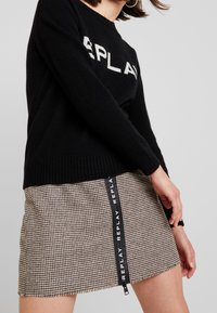 Žena v černém svetr s nápisem "REPLAY" a béžové sukně s houndstooth vzorem, která má černý vertikální zip s detailem "REPLAY".
