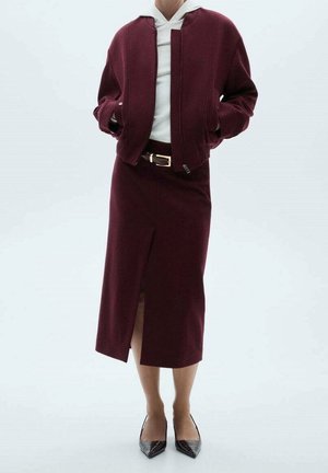 Femme portant une veste bordeaux et une jupe fendue à l'avant, un haut blanc, des escarpins noirs pointus, les mains dans les poches de la veste.