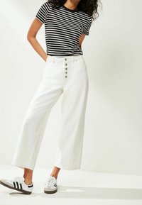 Czarno-biała koszulka w paski, białe spodnie o szerokich nogawkach z wysokim stanem i sześcioma guzikami z przodu, zestawione z czarno-białymi trampkami.