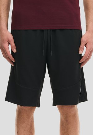 Man draagt zwarte shorts met trekkoord en een donkerrood shirt, staat met ontspannen handen langs zijn zij op een effen lichte achtergrond.