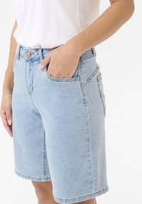 Shorts in denim bluastro con vita alta, tasche frontali e rivetti color beige. Il tessuto ha una consistenza liscia e un design semplice.