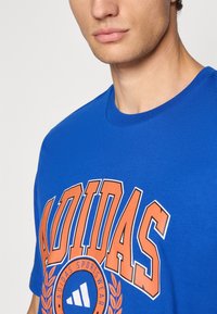 Mėlyna medvilninė marškinėliai su ryškiu oranžiniu užrašu "ADIDAS", papuošti apskritimu logotipu ir lauro akcentais. Trumpomis rankovėmis, apvalia kaklo iškirpte.