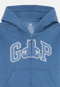 Modrá detská mikina s kapucňou a zipsom, na ktorej sú písmenká "GAP" a dizajn teddy medvedíka integrovaný do loga na hrudi.