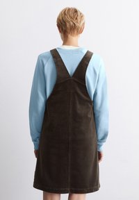 Braunes Cord-Overall-Kleid mit einem taillierten Oberteil, breiten Trägern und einem ausgestellten Saum, darüber getragen über einem hellblauen Langarmshirt.