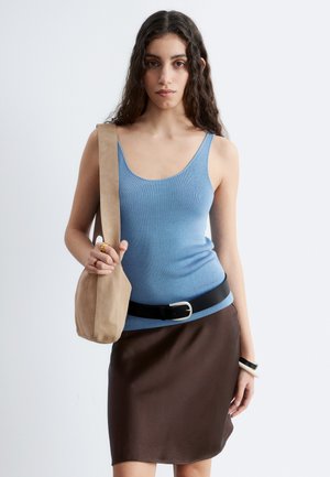 Jeune femme aux longs cheveux foncés portant un débardeur bleu, une jupe marron avec une ceinture noire, tenant un sac à bandoulière beige sur un fond uni.