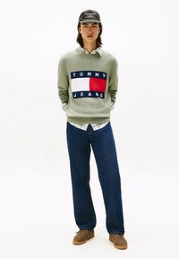 Maglione verde con logo "TOMMY JEANS", camicia a quadri a maniche lunghe sotto, jeans blu e sneakers marroni scuri. Cappellino nero con logo.