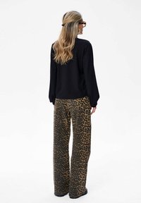 Frau mit langen blonden Haaren in Schildkrötenbrille, die ein schwarzes Langarmoberteil und weit geschnittene Hosen im Leopardenmuster trägt und mit dem Rücken zur Kamera steht.