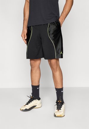 Pantaloncini sportivi neri con piping verde, realizzati in un tessuto sintetico liscio. Indossati con calze nere e sneaker beige.