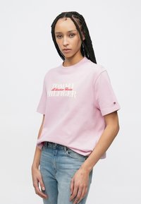 Rosa Baumwoll-T-Shirt mit kurzen Ärmeln. Vorn mit weißem und rotem "TOMMY HILFIGER"-Logo. Kombiniert mit hellblauen Jeans aus Denim.