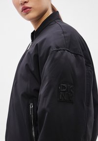 Čierna bomber bunda s hladkou, vypchávkovou textúrou; obsahuje vrecko na zips a čiernu nášivku s logom "DKNY" na ľavom rukáve.