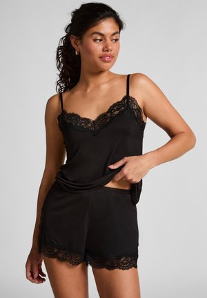 Femme en camisole noire avec dentelle et shorts assortis, levant légèrement le haut, avec des cheveux bouclés attachés et des boucles d'oreilles en or.