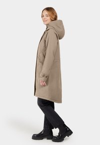 Beige, gewatteerde jas met rechte snit en knoopdetails. Glad stof, zijzakken, gedragen met zwarte jeans en stevige zwarte laarzen.
