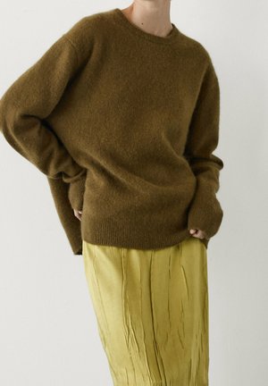Persoon draagt een losse bruine, pluizige trui en een geplooide, lichtgele rok, staand met handen in de mouwen van de trui gestopt.