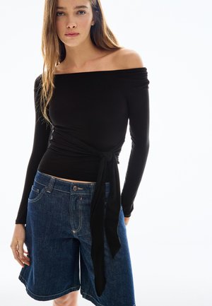 Kobieta w czarnej bluzce z opuszczonymi ramionami, z długimi rękawami i przewiązana w talii, oraz w niebieskich jeansowych szortach do kolan, stojąca na białym tle.