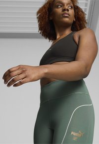 Leggings atletici verdi con vita alta, caratterizzati da cuciture bianche e un logo dorato, abbinati a un reggiseno sportivo nero. Texture liscia.