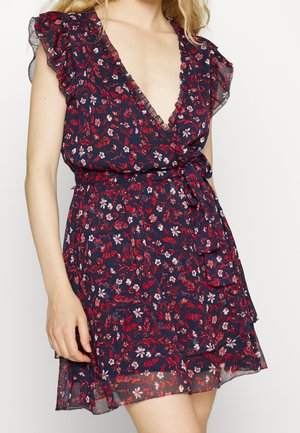 Vrouw draagt een marineblauwe wikkeljurk met een rood-wit bloemmotief, gerimpelde kapmouwen en een strik in de taille.