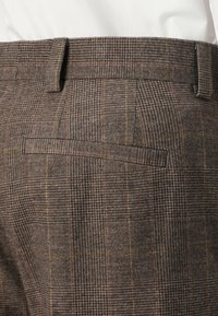 Brown plaid trousers with a textured fabric, featuring a straight waistband and a single back pocket. Subtle tones of yellow and grey in the pattern.