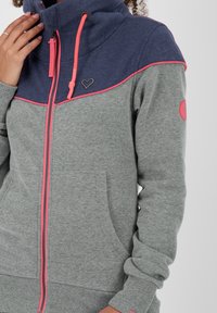 Felpa grigia e blu navy con zip e dettagli rosa, dotata di un alto colletto, tasche frontali e un logo a forma di cuore sul petto. Tessuto morbido e testurizzato.