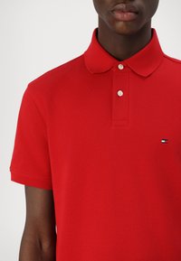 Persona che indossa una polo rossa a maniche corte con chiusura a due bottoni e un piccolo logo Tommy Hilfiger sul petto.