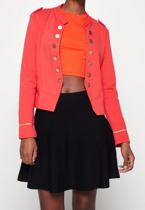 Persoon draagt een feloranje crop top, koraalkleurig militair jasje met gouden knopen en een zwarte uitlopende rok, staand tegen een effen achtergrond.