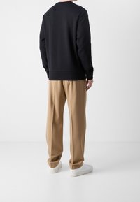 Felpa nera con maniche lunghe e scollatura a girocollo, abbinata a pantaloni beige plissettati e sneakers bianche, vista da dietro.