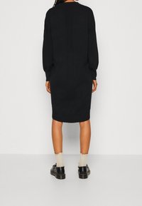 Vestito nero in maglia con maniche lunghe, silhouette dritta e dettagli a coste. Indossato con scarpe nere e calze chiare con bordo smerlato.