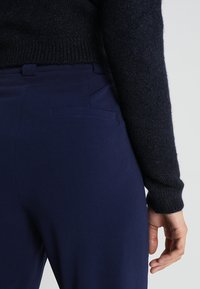 Marineblauwe broeken van gladde stof, met een achterzak en gecombineerd met een gestructureerde zwarte trui. De pasvorm is strakkend en op maat gemaakt.