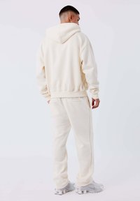 Hombre en sudadera crema y pantalones de chándal a juego se encuentra de espaldas, vistiendo zapatillas deportivas blancas y grises sobre un fondo claro liso.