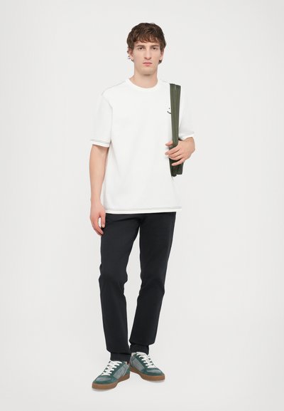 White short-sleeve t-shirt with contrasting seams, paired with dark pants and green suede sneakers with white laces, holding a green item.
