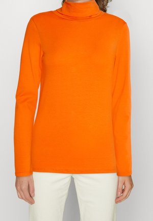 Adulto con top de cuello alto de manga larga de color naranja brillante y pantalones blancos, de pie contra un fondo claro y liso.