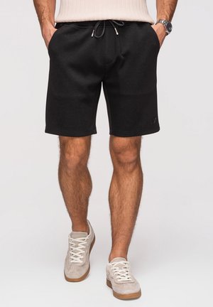 Mann trägt schwarze knielange Shorts mit Kordelzug, beige Sneakers, beige ärmelloses Oberteil und eine Armbanduhr, steht mit den Händen in den Taschen.