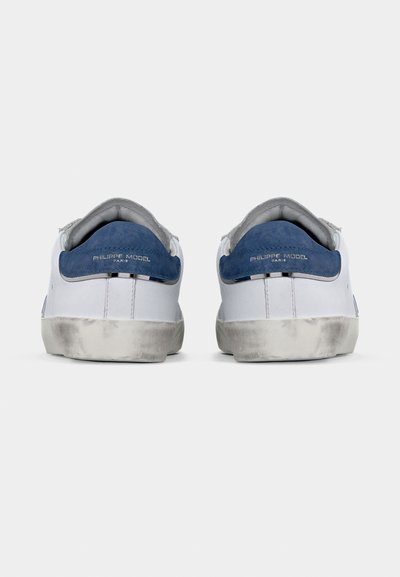 Ζευγάρι λευκά αθλητικά παπούτσια με μπλε ταμπελάκια τακουνιού από σουέτ, λογότυπο "Philippe Model Paris" και φθαρμένες, ελαφρώς βρώμικες σόλες, που φαίνονται από πίσω.