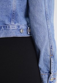 Veste en denim en tissu bleu clair avec un aspect délavé, boutons en métal, coutures visibles et une longueur légèrement raccourcie au niveau de la taille.