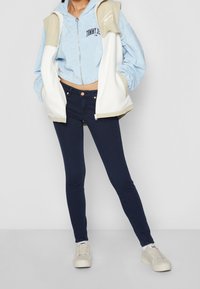 Sudadera con capucha azul de terciopelo recortada con una capa exterior con cremallera blanca y beige, combinada con jeans oscuros ajustados y zapatillas beige.